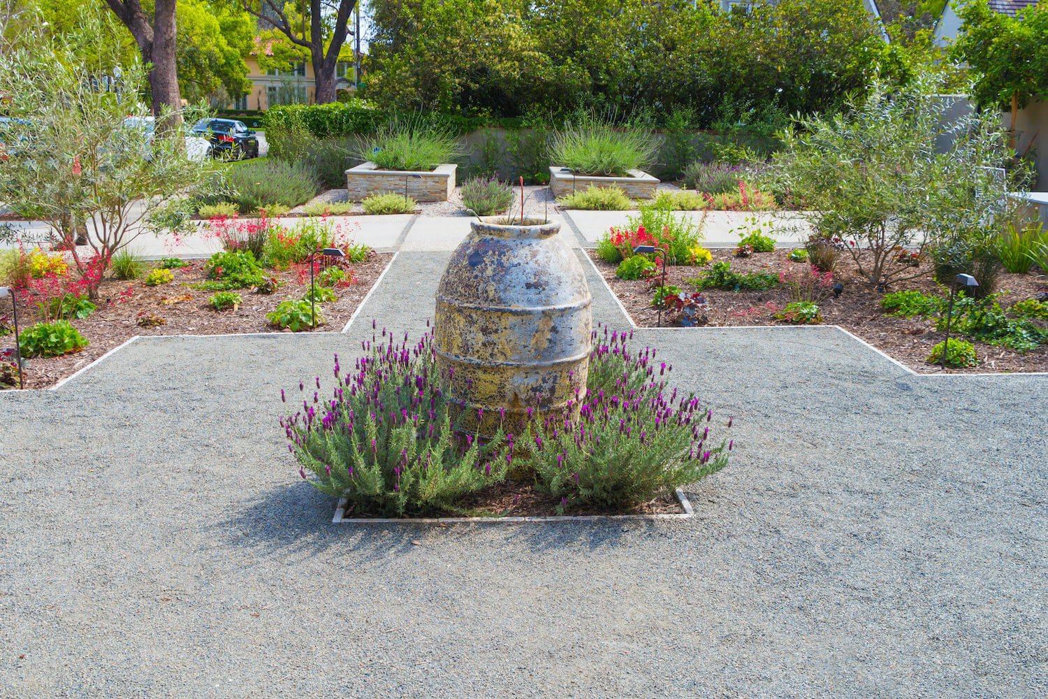 Landscaped courtyard garden with a weathered stone fountain, purple flowers, gravel paths, raised beds, and lush greenery in sunlight.