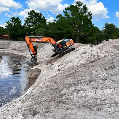 An orange excavator grading a steep, white-sand embankment alongside a body of water.