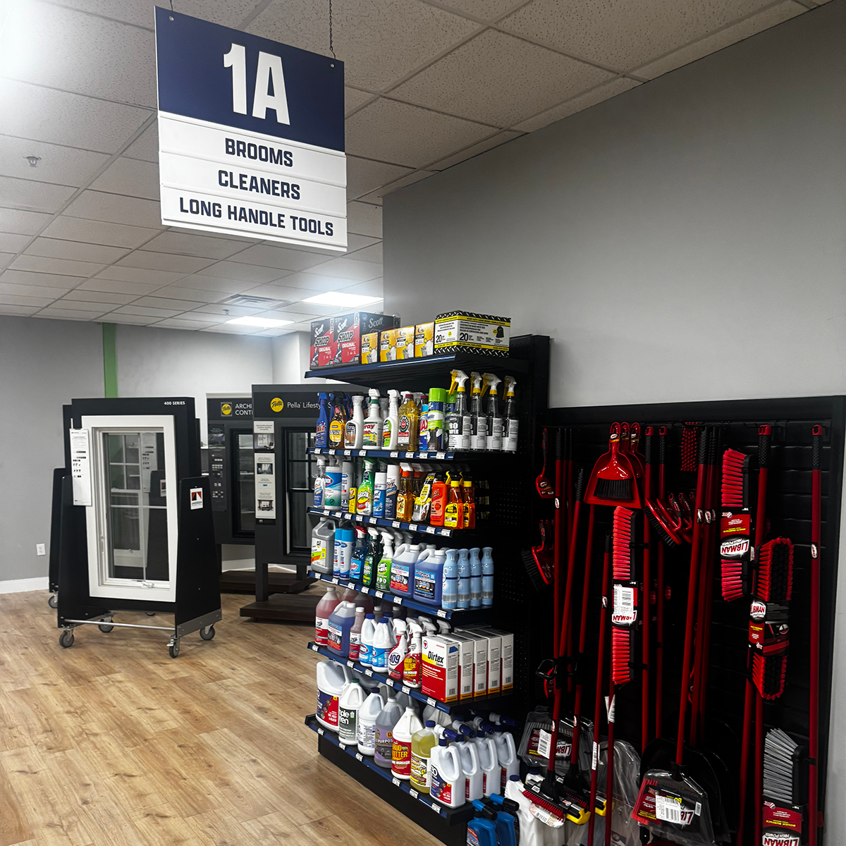 Builders FirstSource store area labeled “1A” for brooms, cleaners and long-handle tools. Shelving displays rows of cleaning sprays, liquid cleaners, paper towels and other household supplies. Red-and-black brooms, brushes and dustpans hang on a black wall at right.