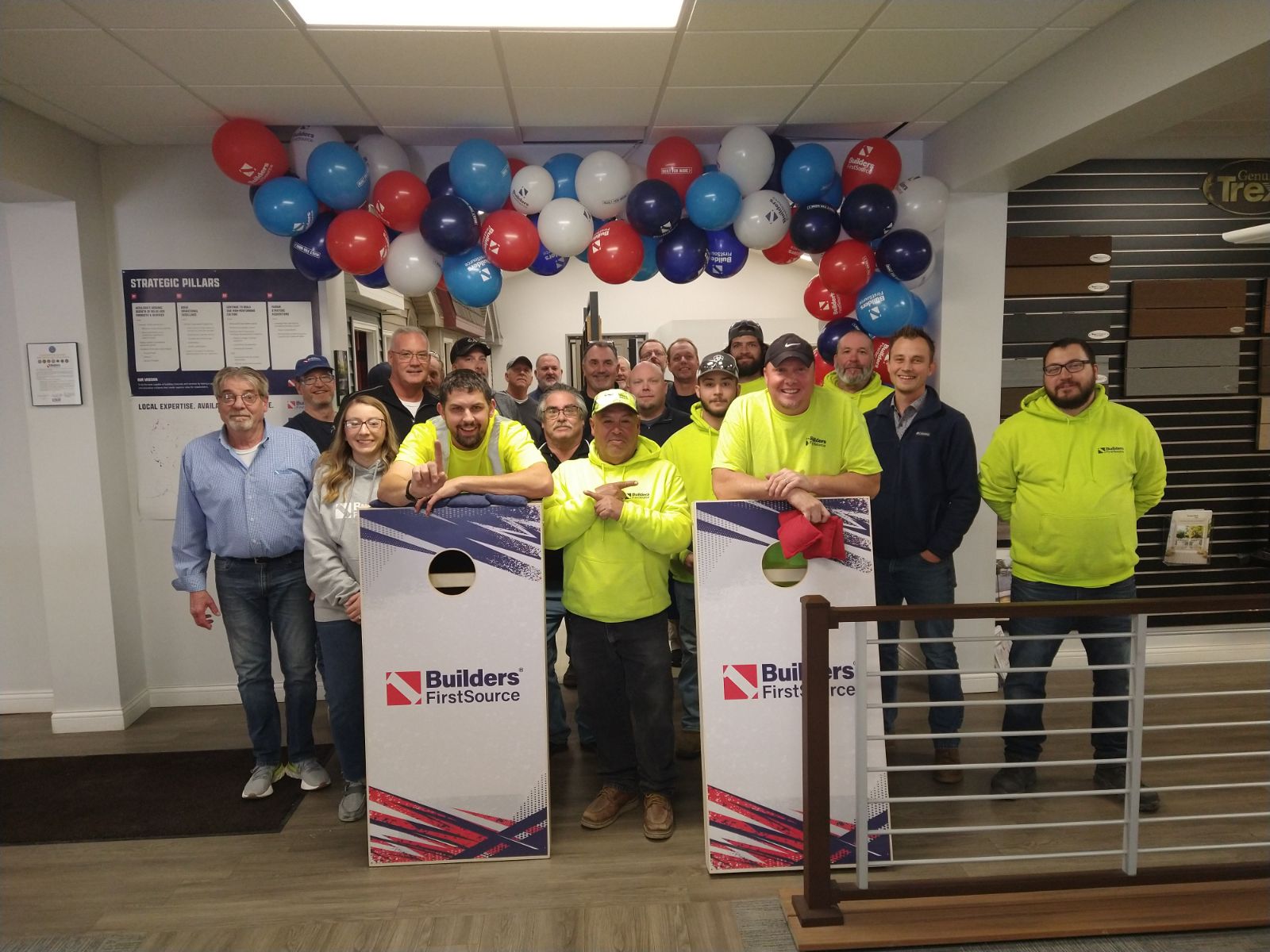 A group photo of the Builders FirstSource team in Fort Wayne, IN. The employees, dressed in vibrant yellow and casual wear, stand under a festive arch of red, white, and blue balloons. A branded cornhole board is displayed prominently in front of the group, symbolizing team spirit and camaraderie. The setting includes a clean, professional office space with informational posters on the walls.