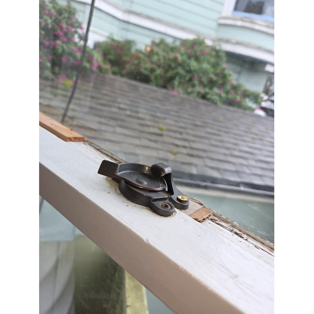 Close-up of a window latch on a painted wooden frame, with chipped paint and a view of a roof and greenery outside.