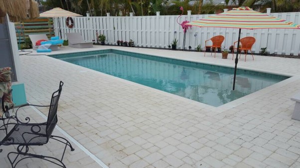 Rectangular backyard swimming pool with light paver decking, patio seating, and a white privacy fence.