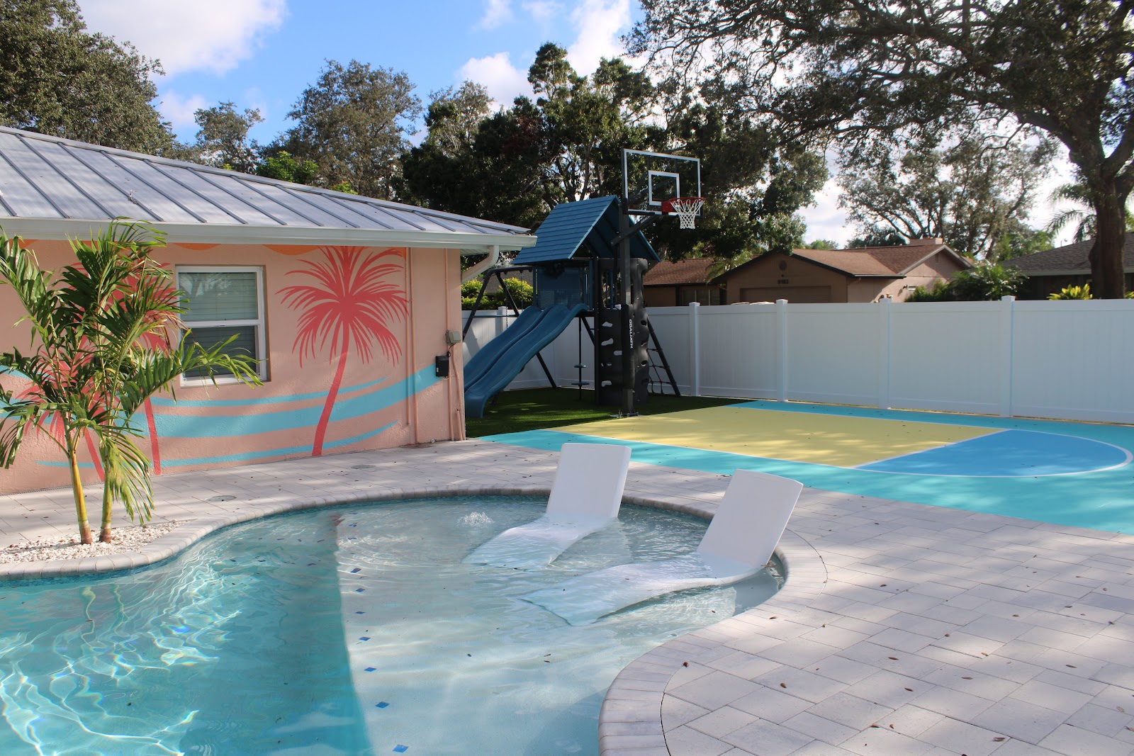 Custom residential pool with a shallow sun shelf, in-pool lounge chairs, colorful patio accents, and a basketball hoop nearby.