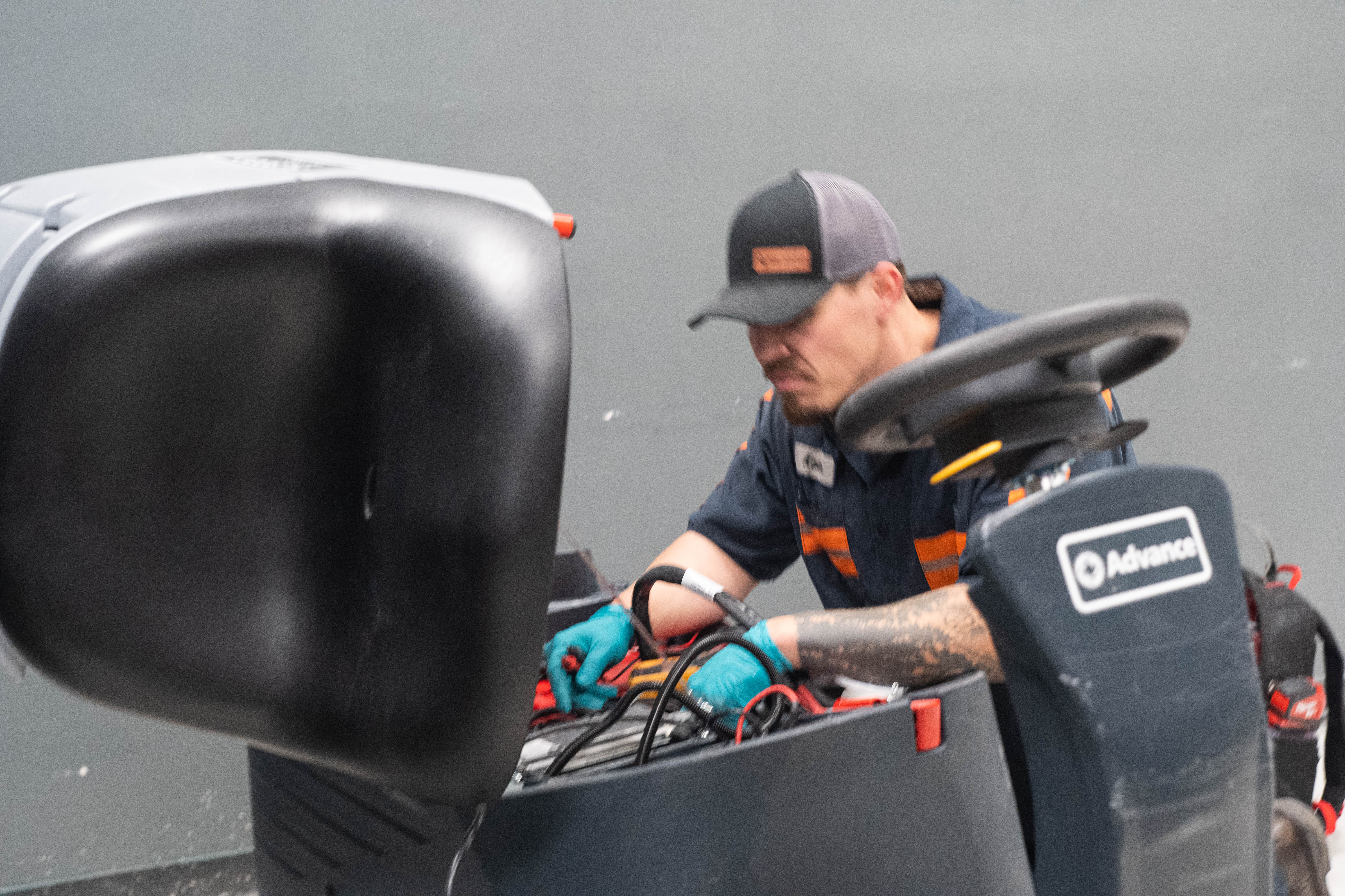 An expert technician from WaySolutions inspects the heavy-duty internal battery bank and electrical wiring of an Advance commercial ride-on floor scrubber. The equipment's upper housing and seat are propped open, allowing the technician to perform a detailed preventive maintenance check on the machine's power systems to ensure maximum runtime.