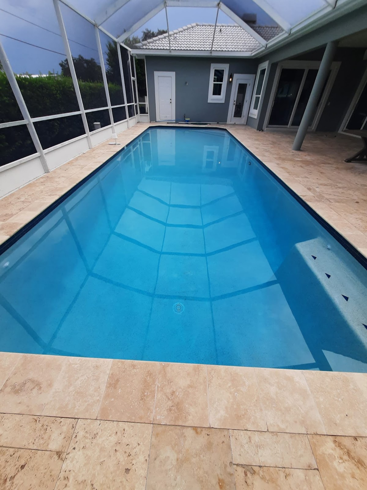 Screen-enclosed rectangular swimming pool with clear blue water, light stone paver decking, and a covered residential patio.