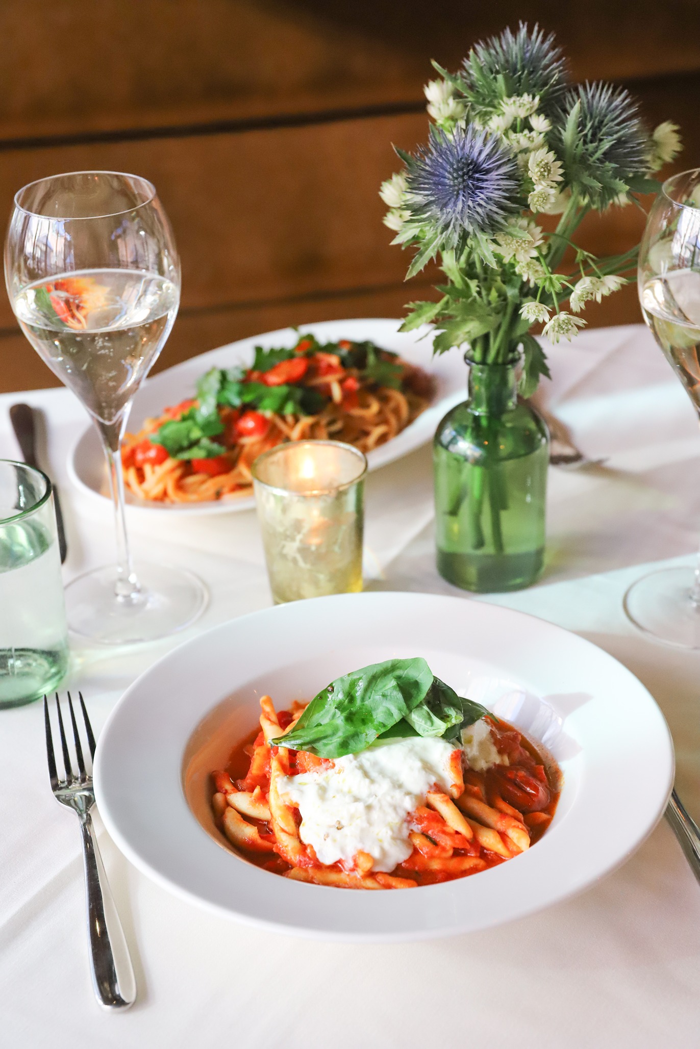 A close-up of a bowl of twisted pasta in a rich red sauce, topped with a generous dollop of creamy burrata and basil. Accompanied by glasses of water and floral decor.