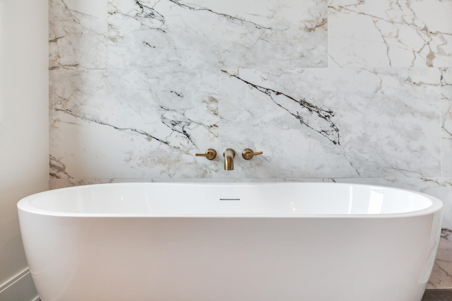 Luxury bathroom detail featuring a freestanding soaking tub, marble-look tile wall, brass fixtures, and a clean, spa-inspired design.