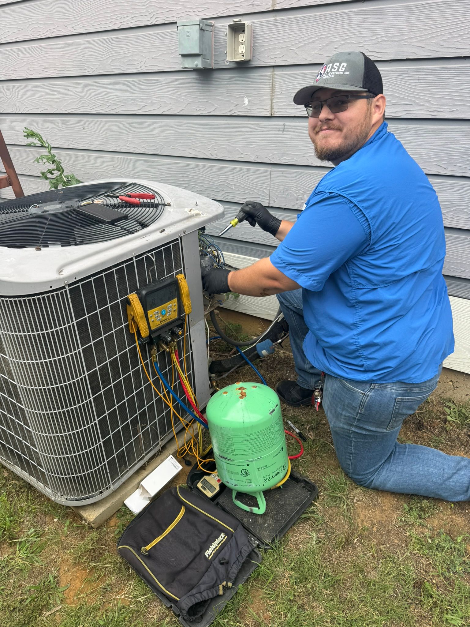 An HVAC technician in a blue shirt and baseball cap is kneeling while servicing an outdoor AC unit. He is using gauges and has a refrigerant tank nearby.
