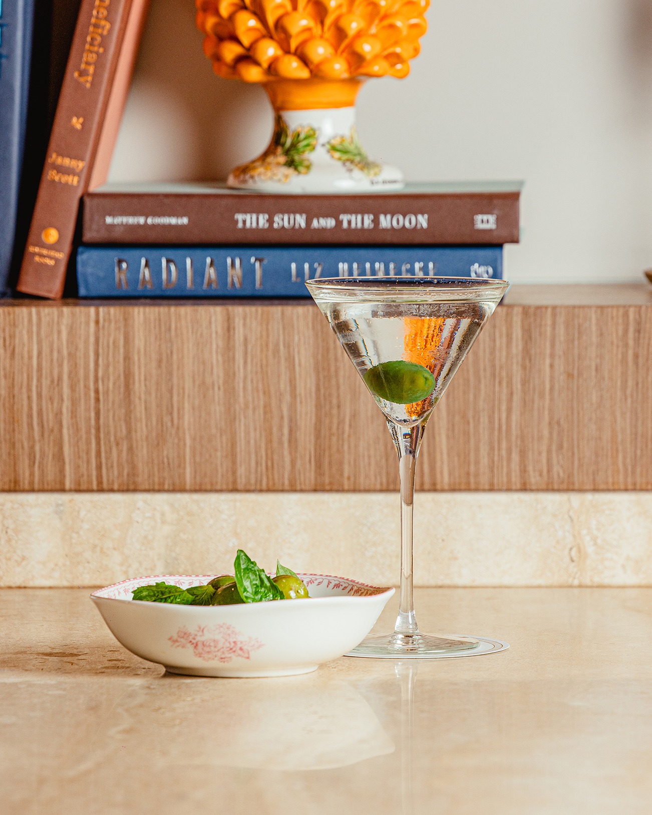 A chilled martini with an olive garnish standing next to a small dish of herb-marinated green olives in front of a curated bookshelf.