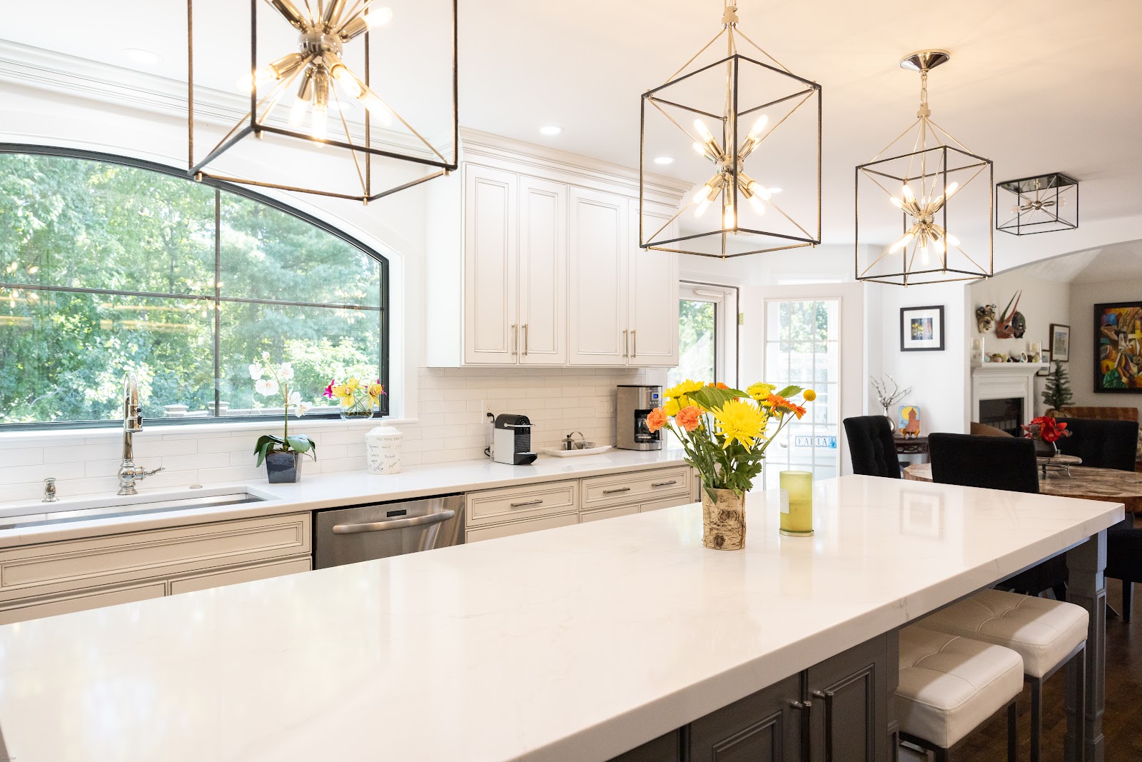 A bright and modern kitchen with white cabinetry, a large arched window overlooking trees, a granite countertop island with bar stools, and contemporary pendant lighting. A bouquet of yellow and orange flowers sits on the island next to a green candle.