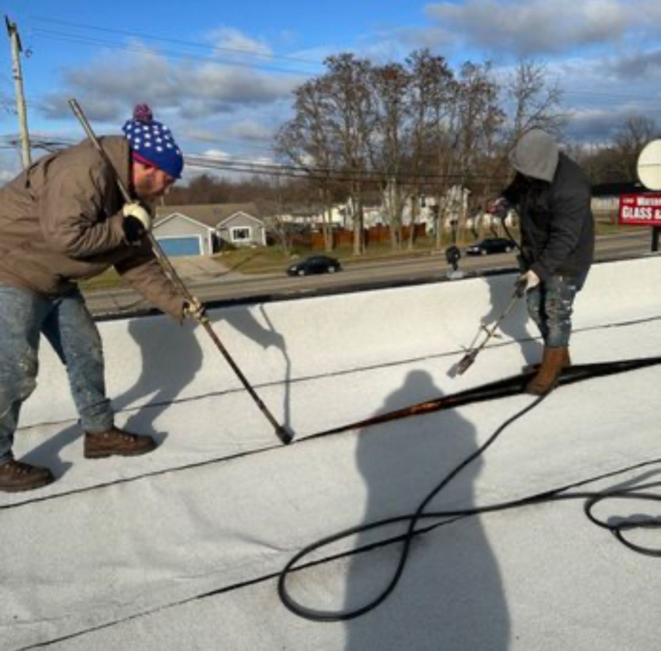 Two workers collaborate on installing a white membrane roof. One applies heat with a torch or heat gun to weld a seam, while the other uses a long-handled tool nearby.