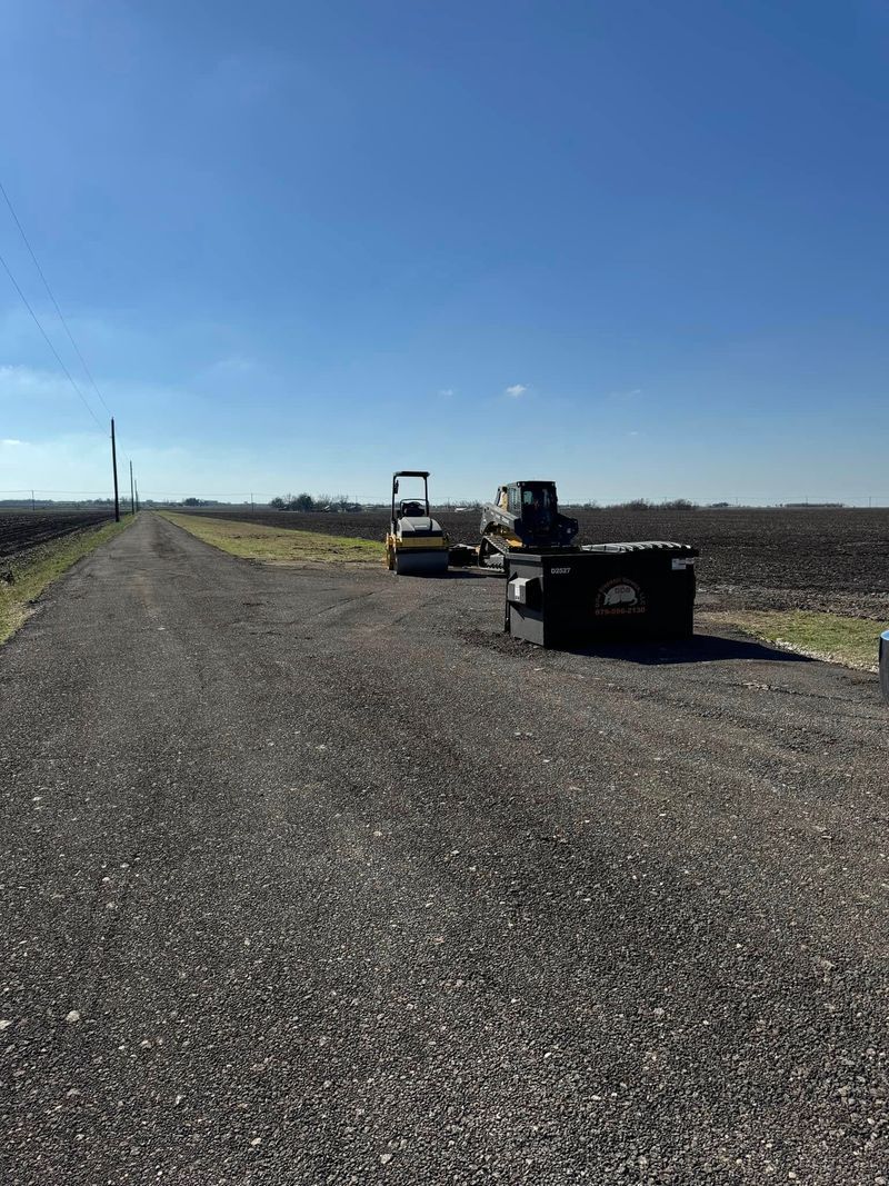 Road construction scene with roller compactor and skid steer positioned on paved rural road, illustrating asphalt work, compaction, and site preparation for infrastructure or driveway development in open landscape setting.