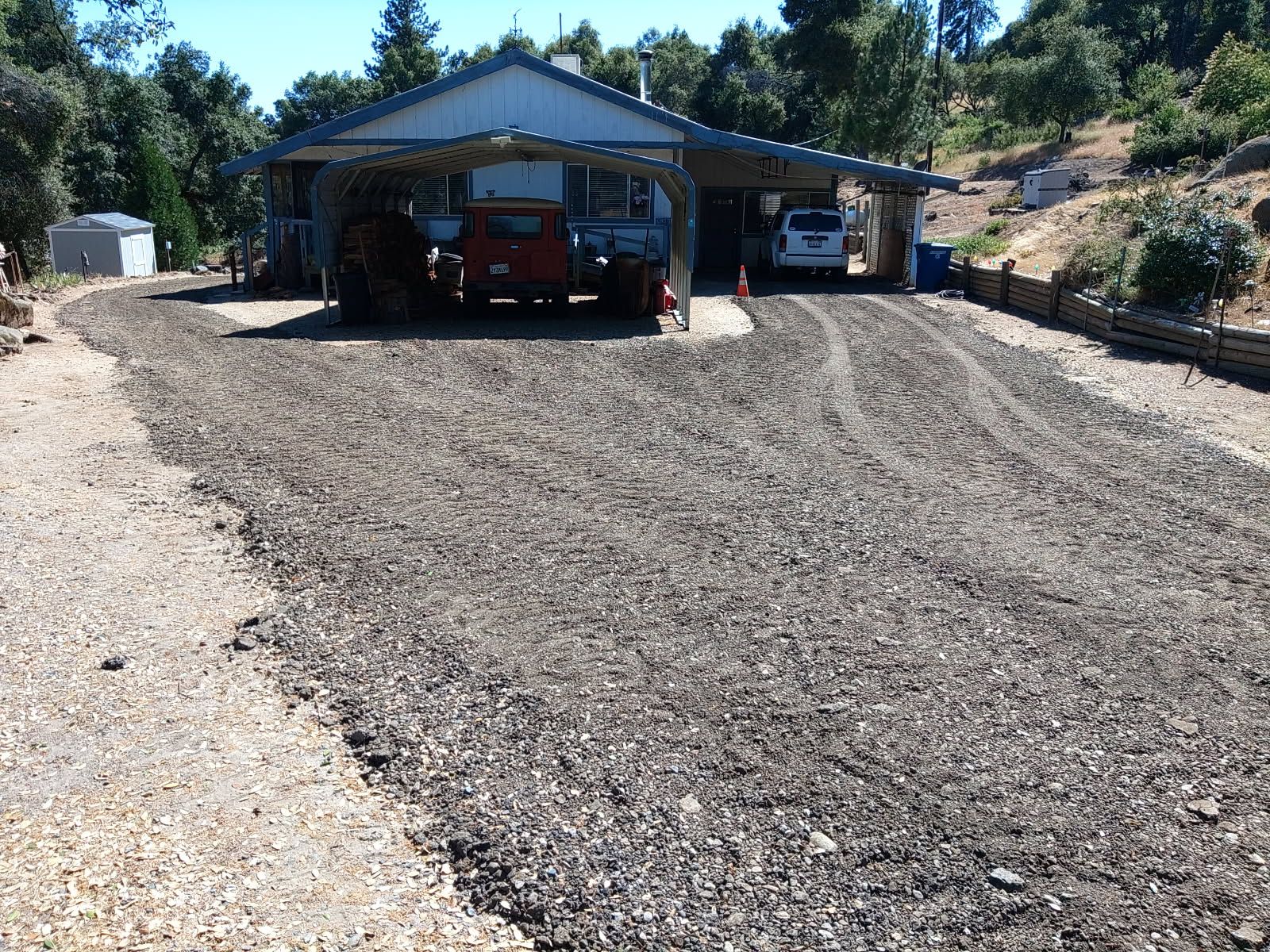 Residential property features a newly spread gravel driveway and parking area leading toward a house and carports, with parked vehicles, retaining structures, trees, hillside vegetation, and outbuildings surrounding the site.