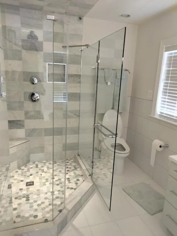 A modern bathroom features a neo-angle shower with glass doors, a built-in bench, and a hexagonal tile floor. Adjacent to the shower is a white toilet with a chrome grab bar and a toilet paper holder on the tiled wall. A light-colored bath mat rests on the white tiled floor, and a window with blinds is visible on the right.