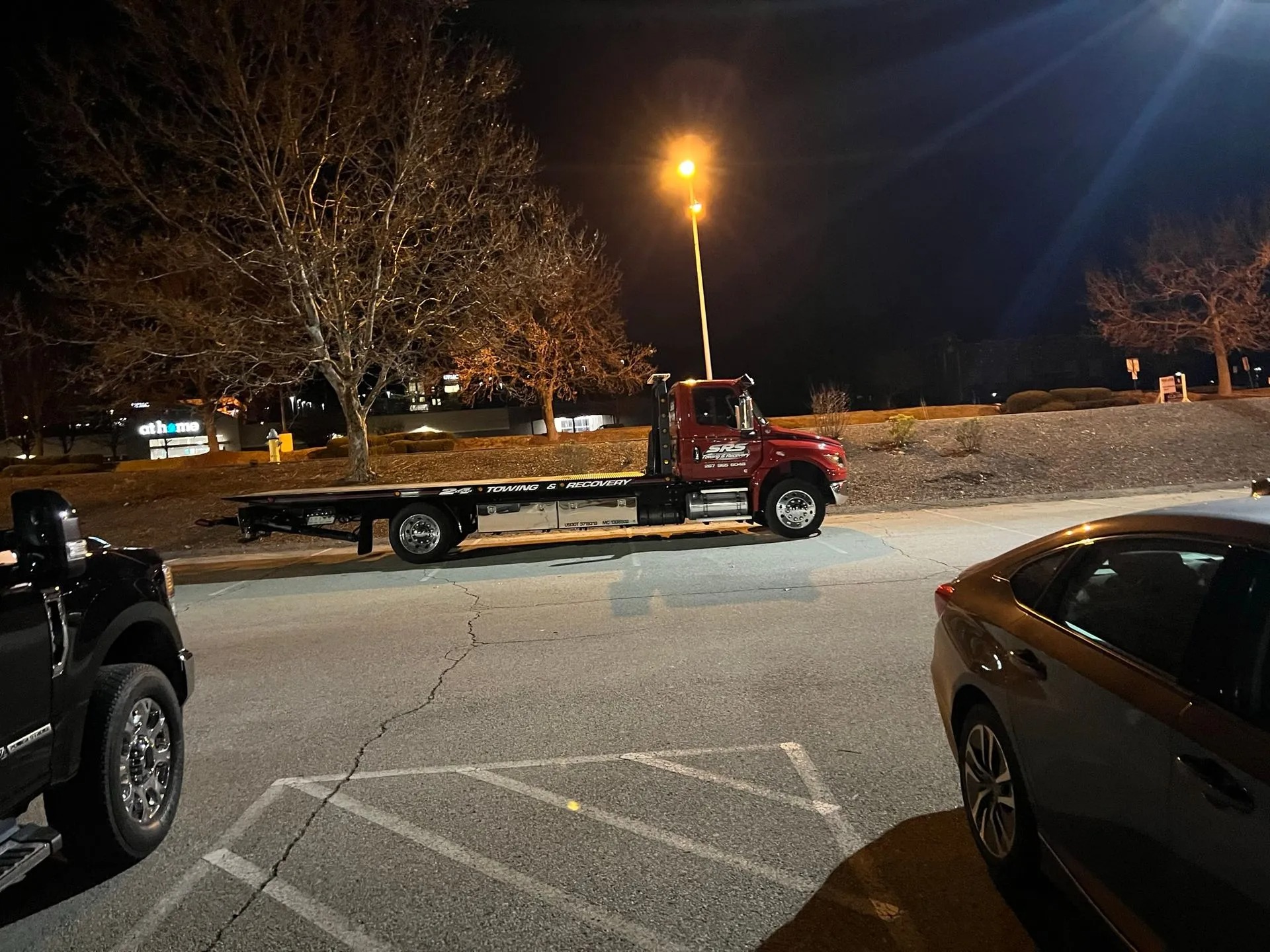 A red SRS Towing & Recovery flatbed tow truck parked alone in a dimly lit parking lot at night, illuminated by streetlights.