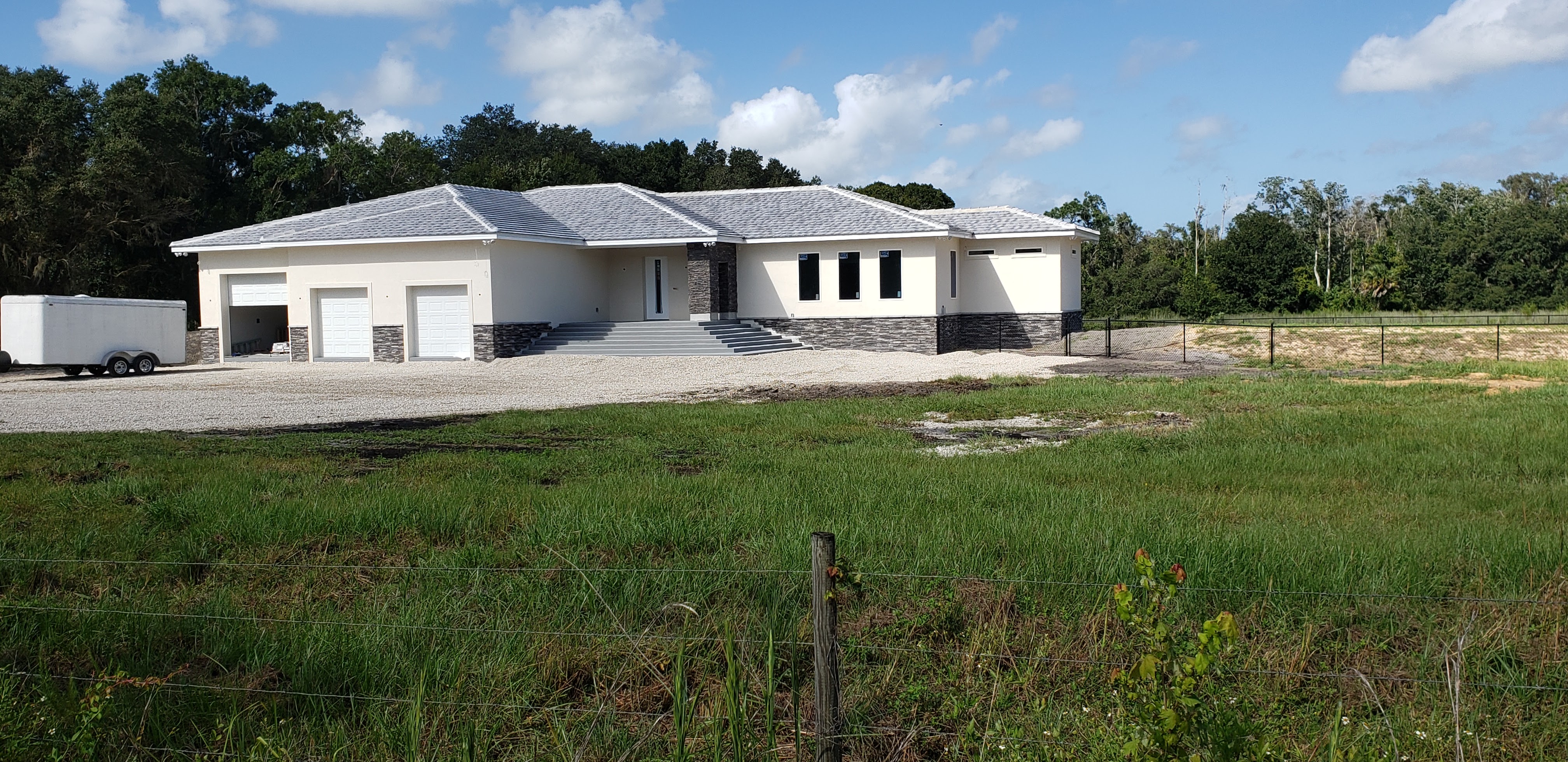 A newly constructed house with a light beige exterior and a grey shingled roof sits on a gravel driv 