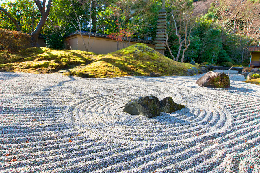 Serene Japanese zen garden with raked white gravel ripples around dark rocks, moss-covered mounds, trees, and a traditional wall.