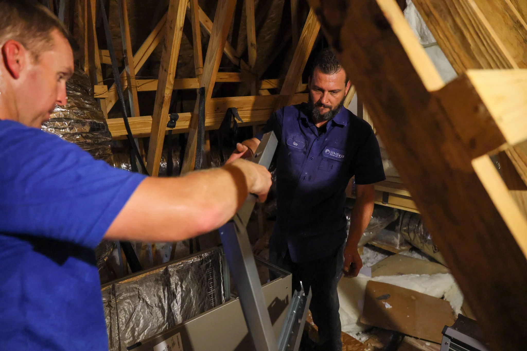 Two men are working in an attic, installing or repairing an HVAC unit. The attic has exposed wooden  