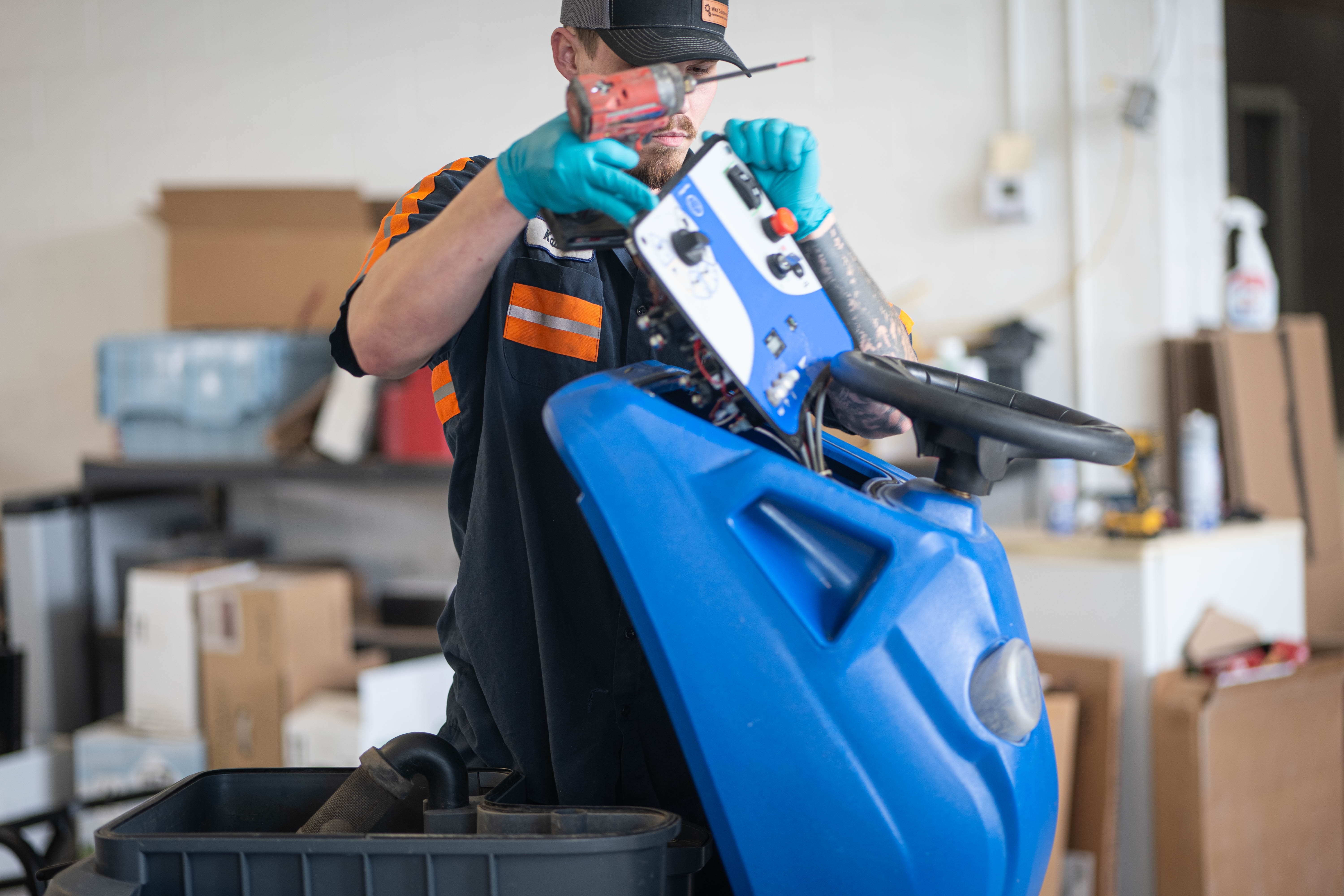 WaySolutions lead technician Kaden utilizes a power drill to carefully disassemble the upper control panel of a blue Kärcher Chariot stand-on commercial floor scrubber. By performing expert top-level electrical and mechanical diagnostics in Layton, Utah, our team ensures that complex supermarket and retail cleaning fleets operate smoothly without causing facility downtime.