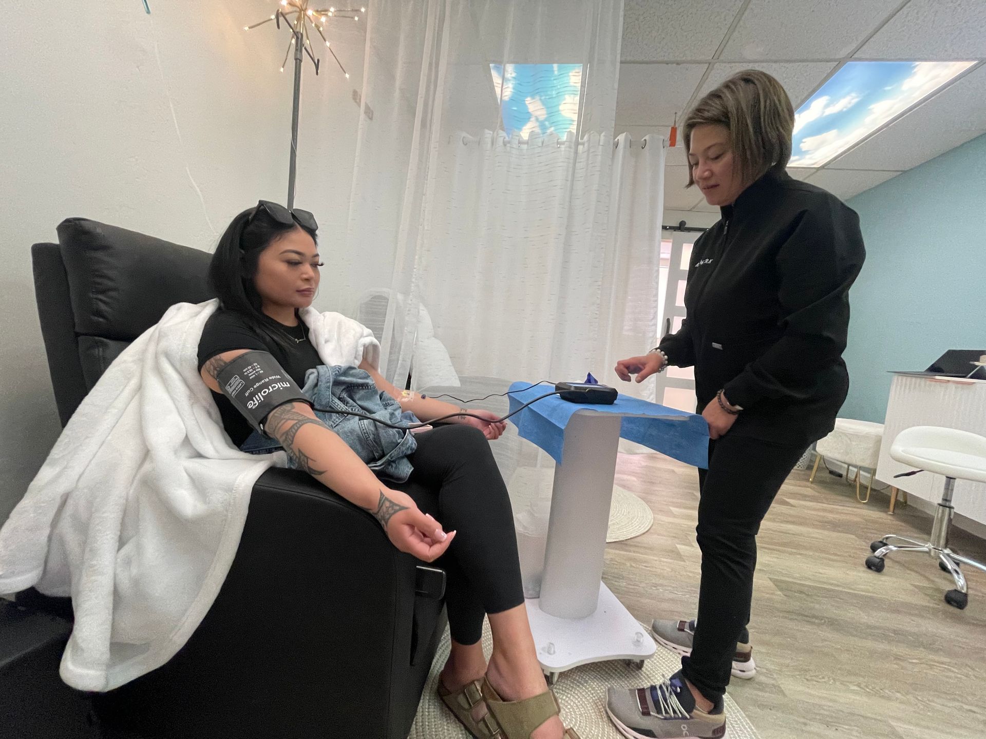 A woman relaxes in a recliner receiving an IV therapy session while a healthcare professional monitors equipment in a calm, modern treatment room with soft lighting, privacy curtains, and comfortable furnishings.