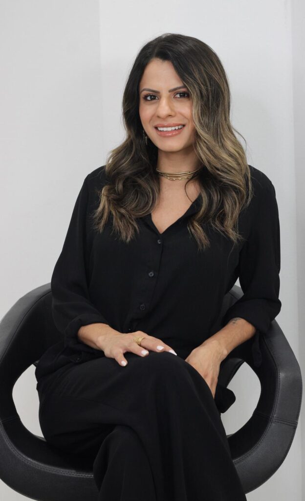 A woman with long, wavy brown hair and a warm smile is seated in a modern black chair against a plain white background. She is wearing a black button-down shirt, black pants, and gold jewelry, including a multi-layered necklace and several rings on her fingers. Her hands are resting on her lap, and her fingernails are painted white. The lighting is bright and even, highlighting her features and the details of her attire.