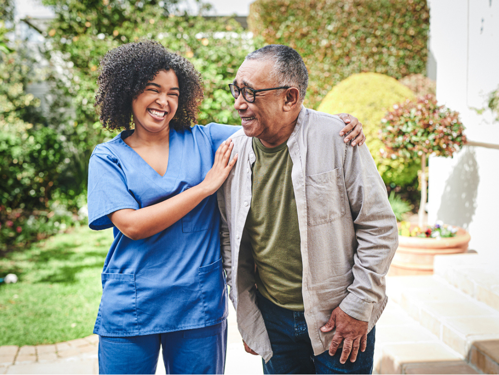 A caregiver standing alongside and supporting a smiling senior man outdoors, representing compassionate personal care, companionship, and daily living assistance.