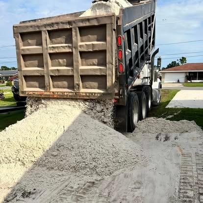 A dump truck with a raised bed unloading a large pile of white sand onto the ground near a residential driveway.
