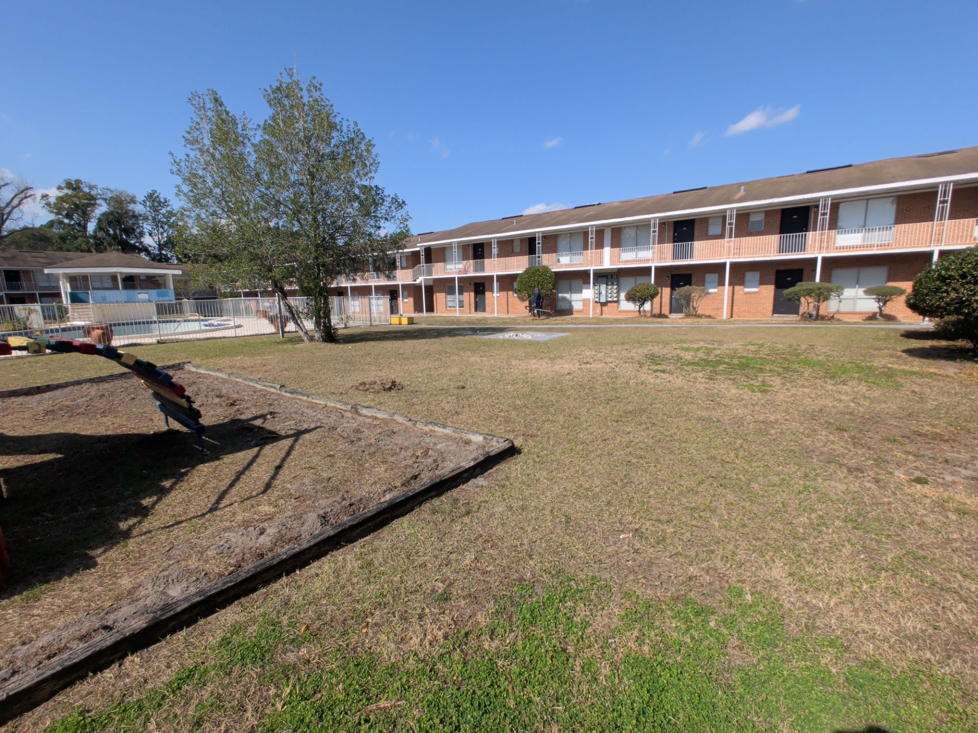 Apartment complex courtyard with grassy lawn, small playground equipment, and fenced swimming pool. Two-story brick residential buildings with balconies surround the shared outdoor community space.