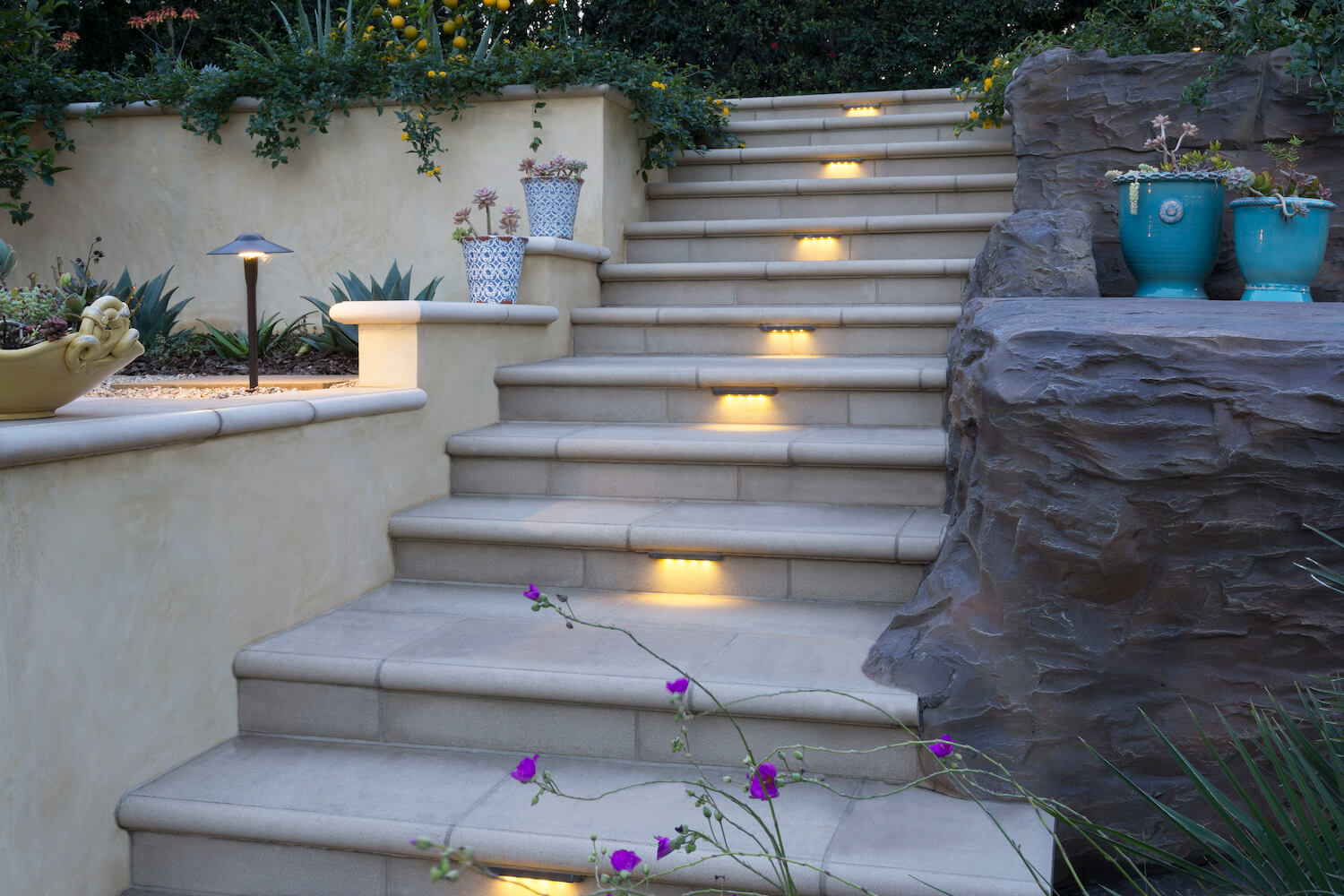 Stone garden staircase with warm step lights, purple flowers, turquoise pots, rock wall, and lush greenery creates a tranquil evening retreat.
