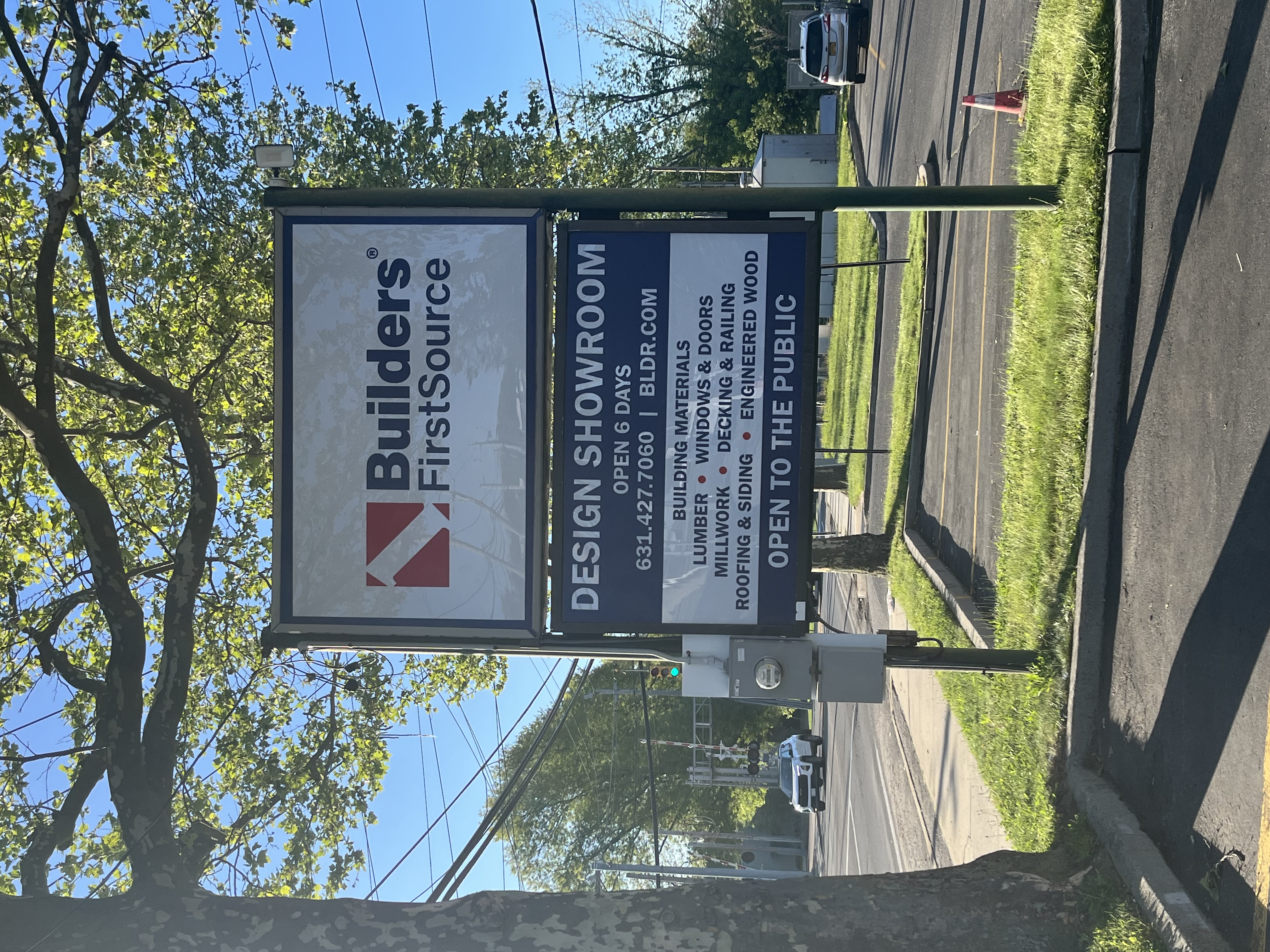 A Builders FirstSource road sign listing showroom hours, contact information, and services like lumber, windows, doors, and millwork with trees and roadway in the background.