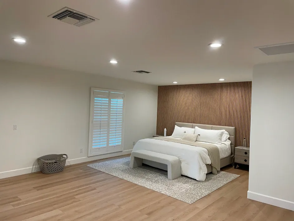Minimalist bedroom with wood flooring, neutral tones, upholstered bed, bench seating, and vertical slat accent wall, complemented by recessed lighting and plantation shutters, creating a calm, modern, and spacious interior environment.