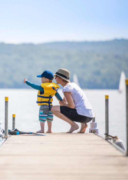 A child in a yellow life jacket and blue cap stands on a wooden dock pointing at a boat, with an adult beside them. A calm lake and hills in the background.