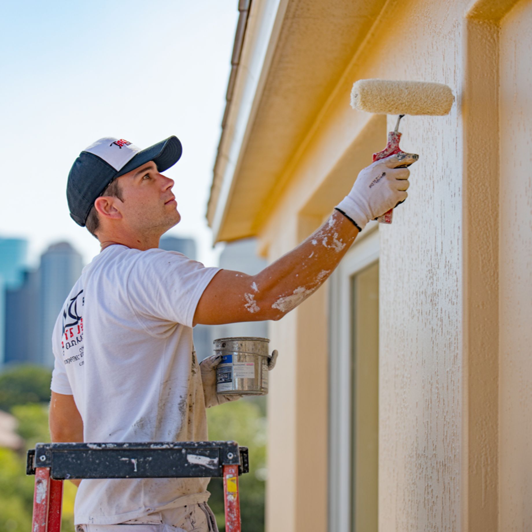 A painter in a white t-shirt and a dark baseball cap with a white front is painting the side of a bu 
