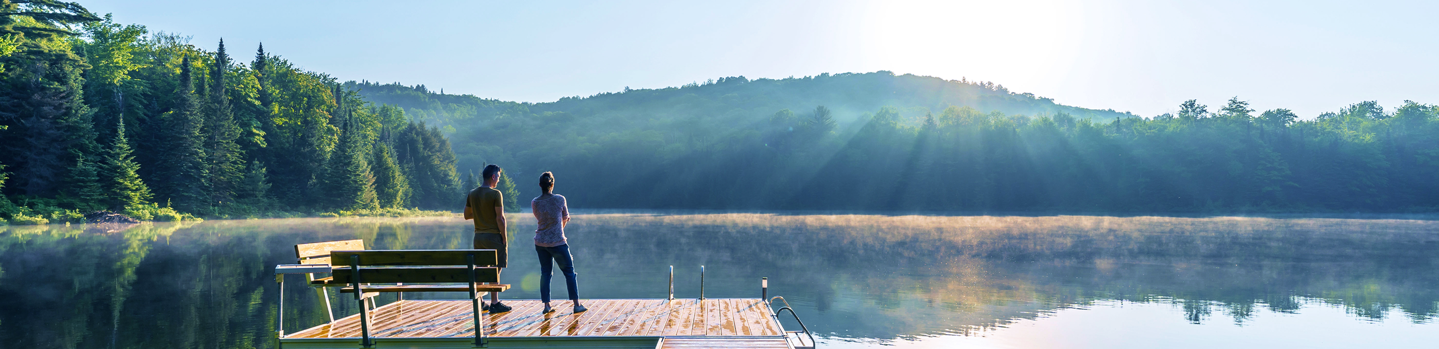 Two individuals standing on a dock overlooking a calm lake at golden hour.
