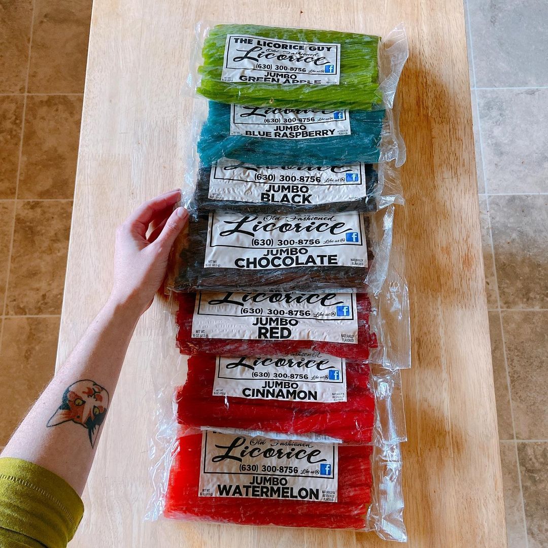 A customer display of The Licorice Guy's seven licorice flavors stacked on a countertop, featuring Green Apple, Blue Raspberry, Black, Chocolate, Red, Cinnamon, and Watermelon varieties, each in a one-pound bag.