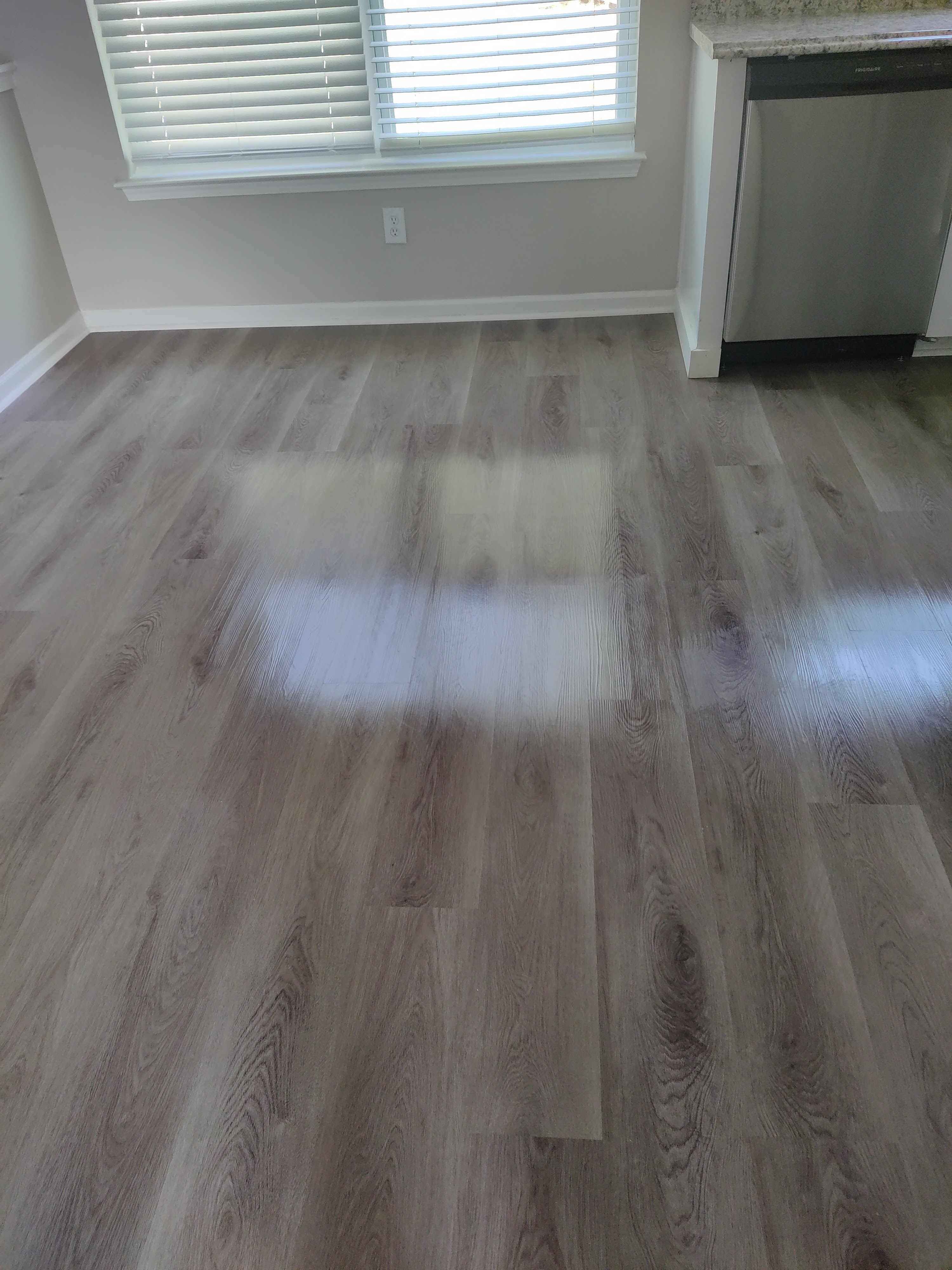 A medium shot shows the floor of a room with a dishwasher in the background. The floor is made of wood-look laminate planks in a gray-brown color. Sunlight is reflecting off the floor near a window with white blinds. The dishwasher is stainless steel and has the "FRIGIDAIRE" logo on the front. A granite countertop is visible above the dishwasher.
