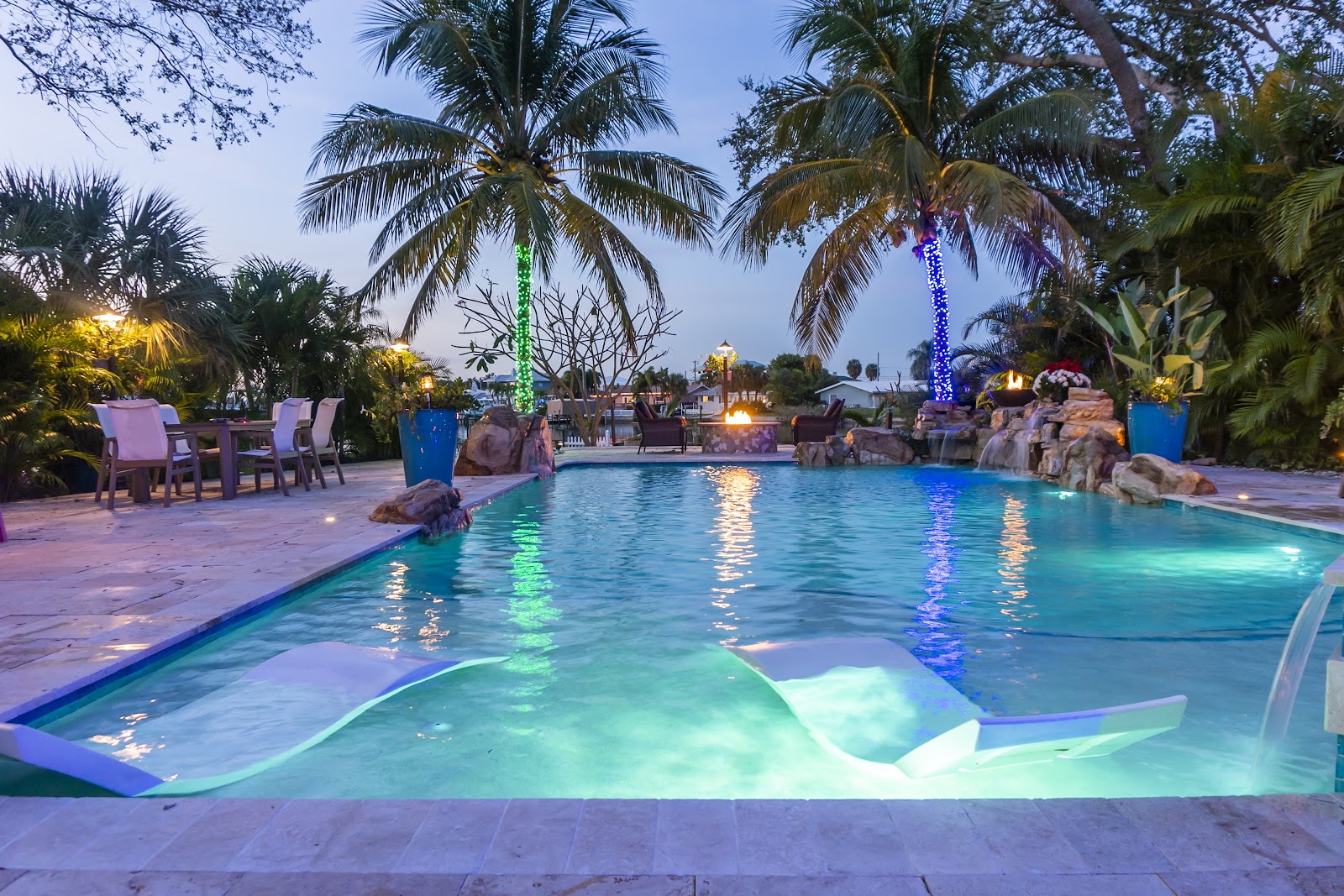 Luxury backyard swimming pool glowing with colorful LED lighting, surrounded by palm trees and tropical landscaping at dusk.