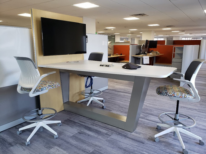 A collaborative workspace featuring a modern high-top table with a sleek white surface and a metallic base. The setup includes four adjustable swivel chairs with patterned seat cushions, adding a touch of color and comfort. A wall-mounted flat-screen TV is integrated into the design, perfect for presentations or video conferencing. The workspace is part of an open office layout, with cubicles and desks visible in the background.