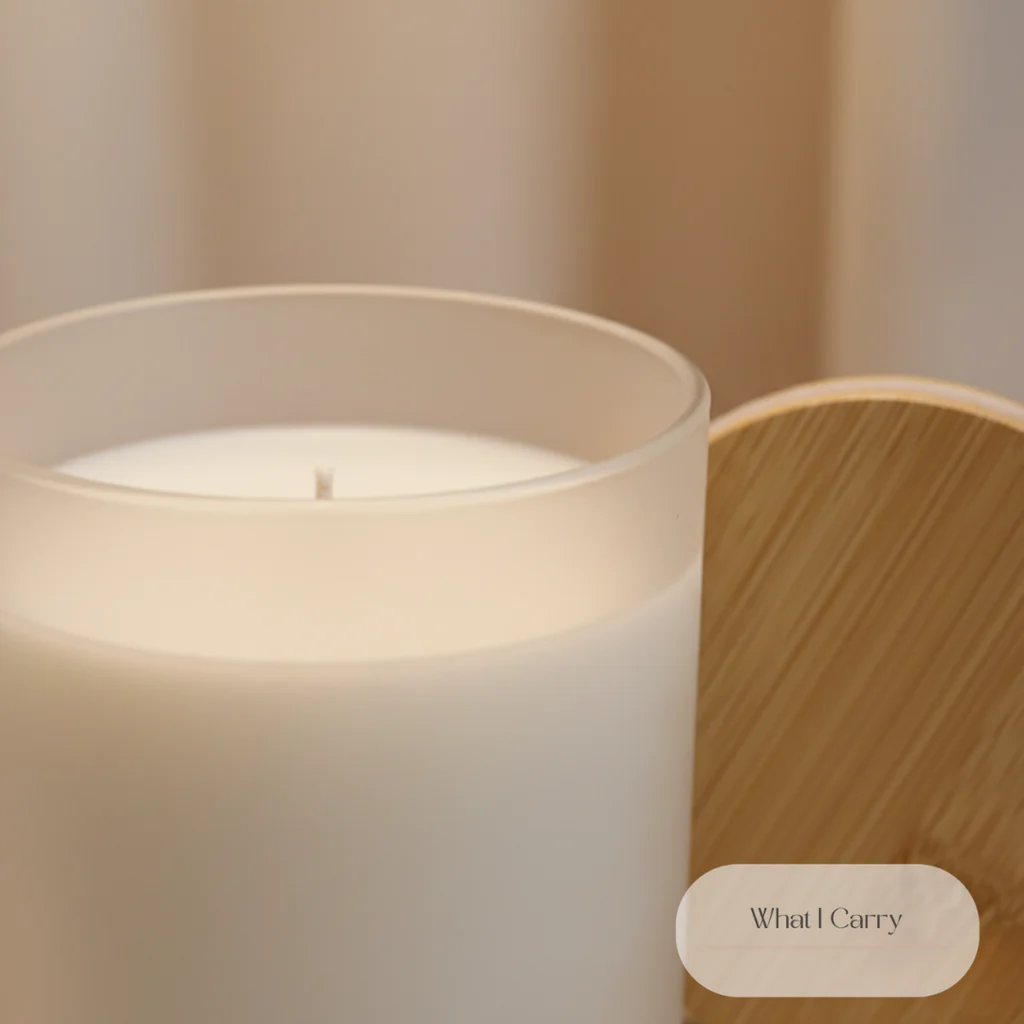 A close-up shot of a white candle in a frosted glass container, next to a round wooden lid with a subtle grain pattern. The background is softly blurred in a light beige color. A small, oval label with the text "What I Carry" is visible in the lower right corner.