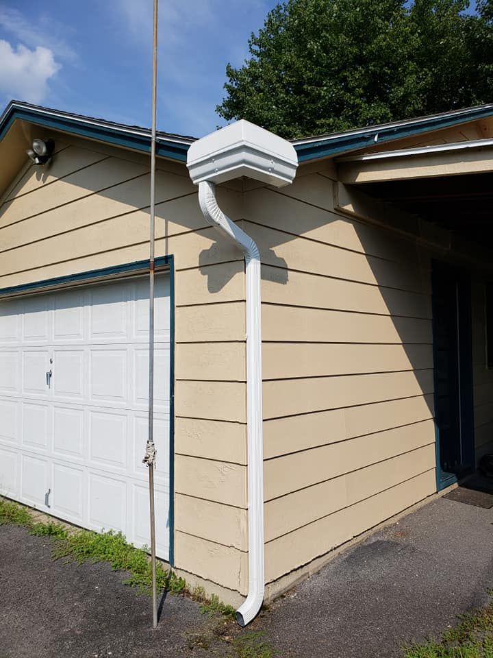 A residential garage corner with beige siding, featuring a white gutter system and downspout connected to a decorative rain head, installed for efficient water drainage beside a driveway.