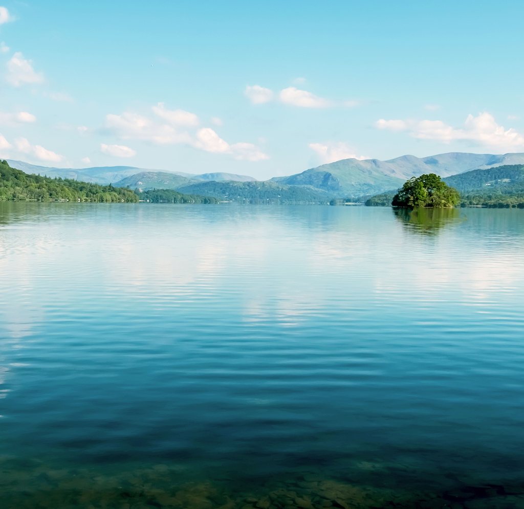 Calm, still waters on a blue lake underneath an equally blue sky with mountains in the distance.