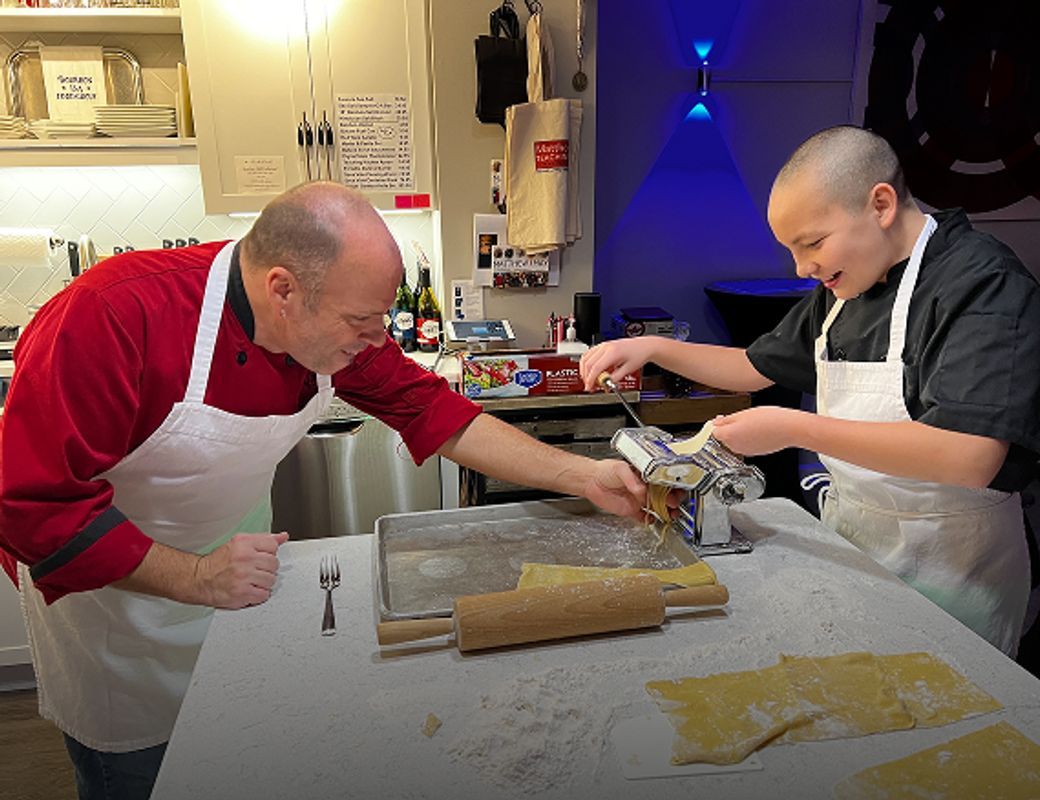 A bald man in a red chef's jacket and a boy in a black chef's jacket are making pasta using a pasta maker. Both are wearing white aprons. The man is leaning over a counter, assisting the boy with the pasta maker. The boy is smiling and holding a sheet of pasta dough as it goes through the machine. Freshly made pasta sheets are on the counter, along with a rolling pin and a fork. The background shows a kitchen with cabinets, shelves stocked with dishes, and a distinctive blue light fixture on the wall.