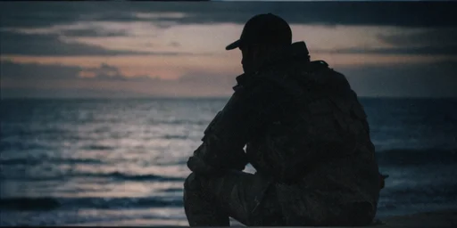 A silhouette of a person wearing a baseball cap and tactical gear sits facing the ocean at dusk, with calm water and a cloudy sky.