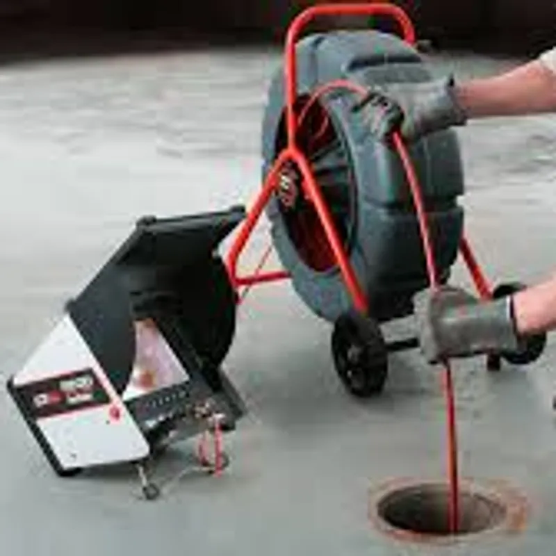 A close-up view of a sewer inspection camera system, with a cable being fed into a manhole. The system includes a large reel with a red frame and a monitor unit with a screen and controls. Two gloved hands are visible, one holding the cable and the other near the reel.