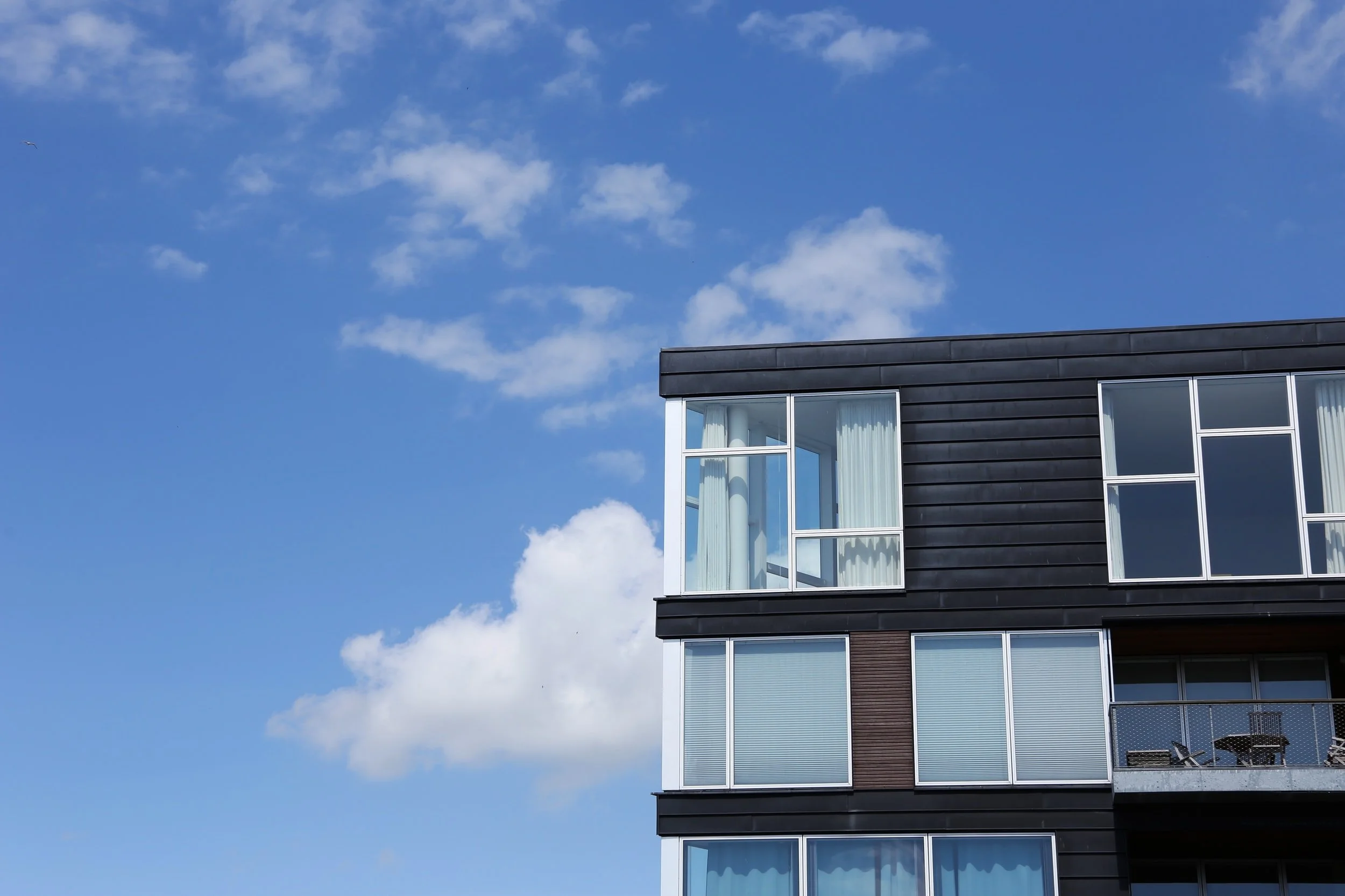 A modern building with dark siding and large windows under a bright blue sky with scattered white clouds. A small bird is visible flying in the distance.