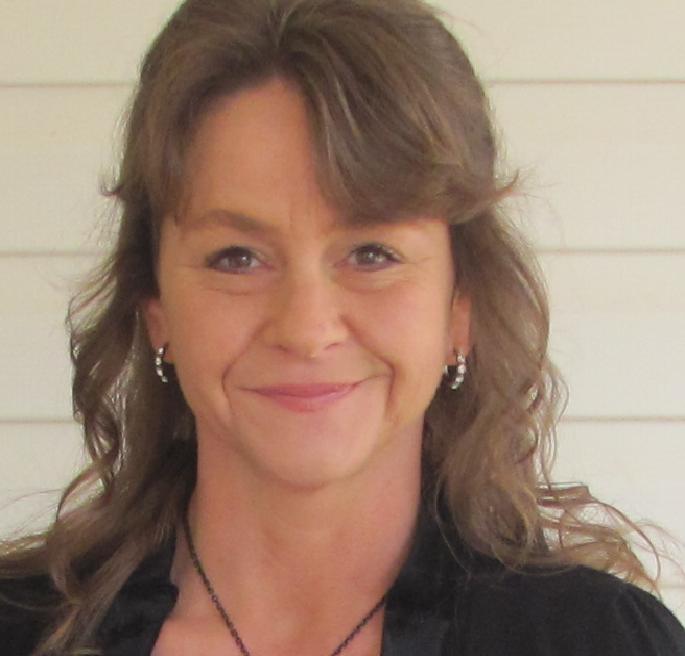 A medium shot of a woman with shoulder-length brown hair smiling. She is wearing a black top and a necklace. The background is a light-colored wall with horizontal lines.