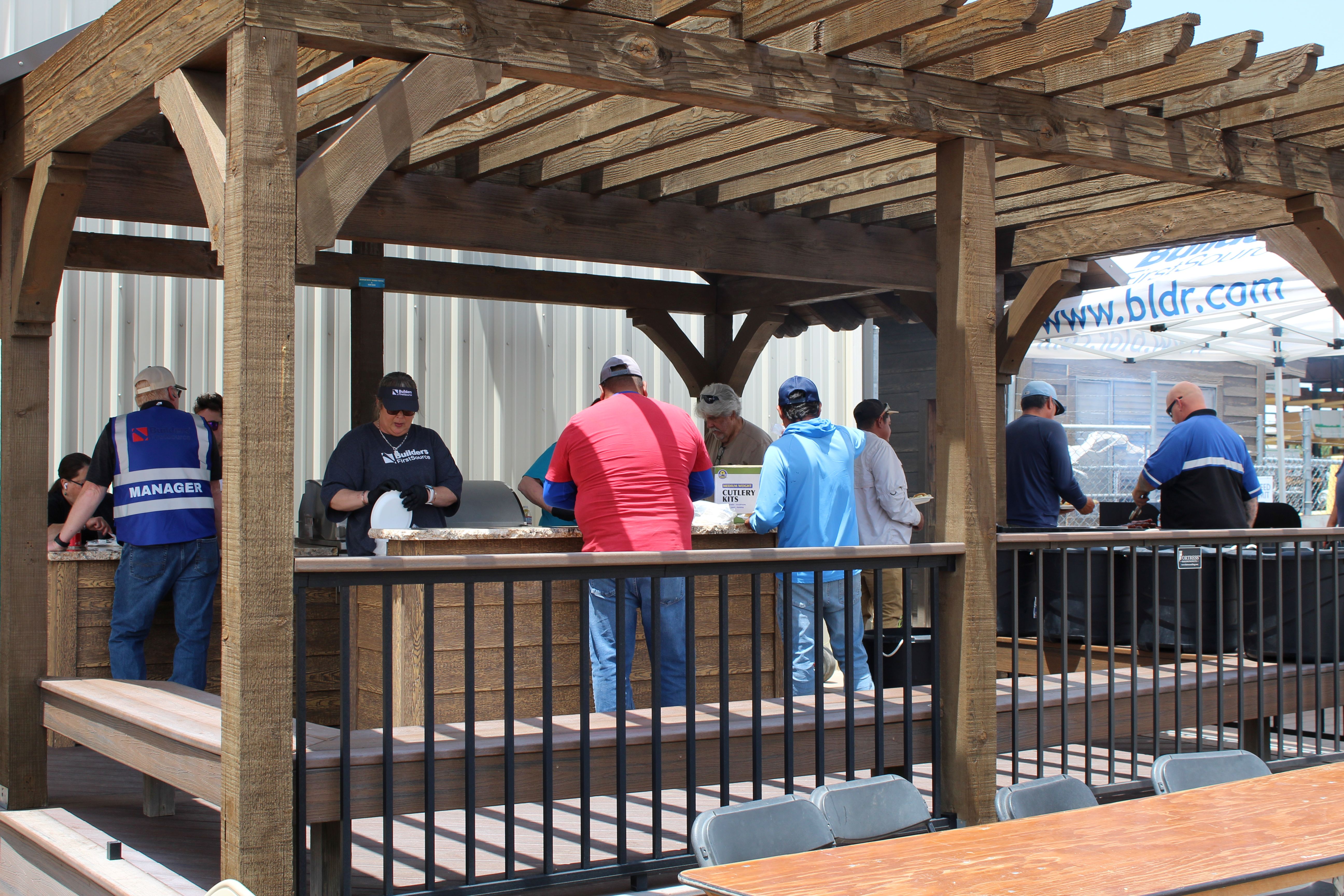 Customers and team members gather under a timber pergola at a Builders FirstSource customer appreciation event, enjoying food and conversation around an outdoor kitchen display.