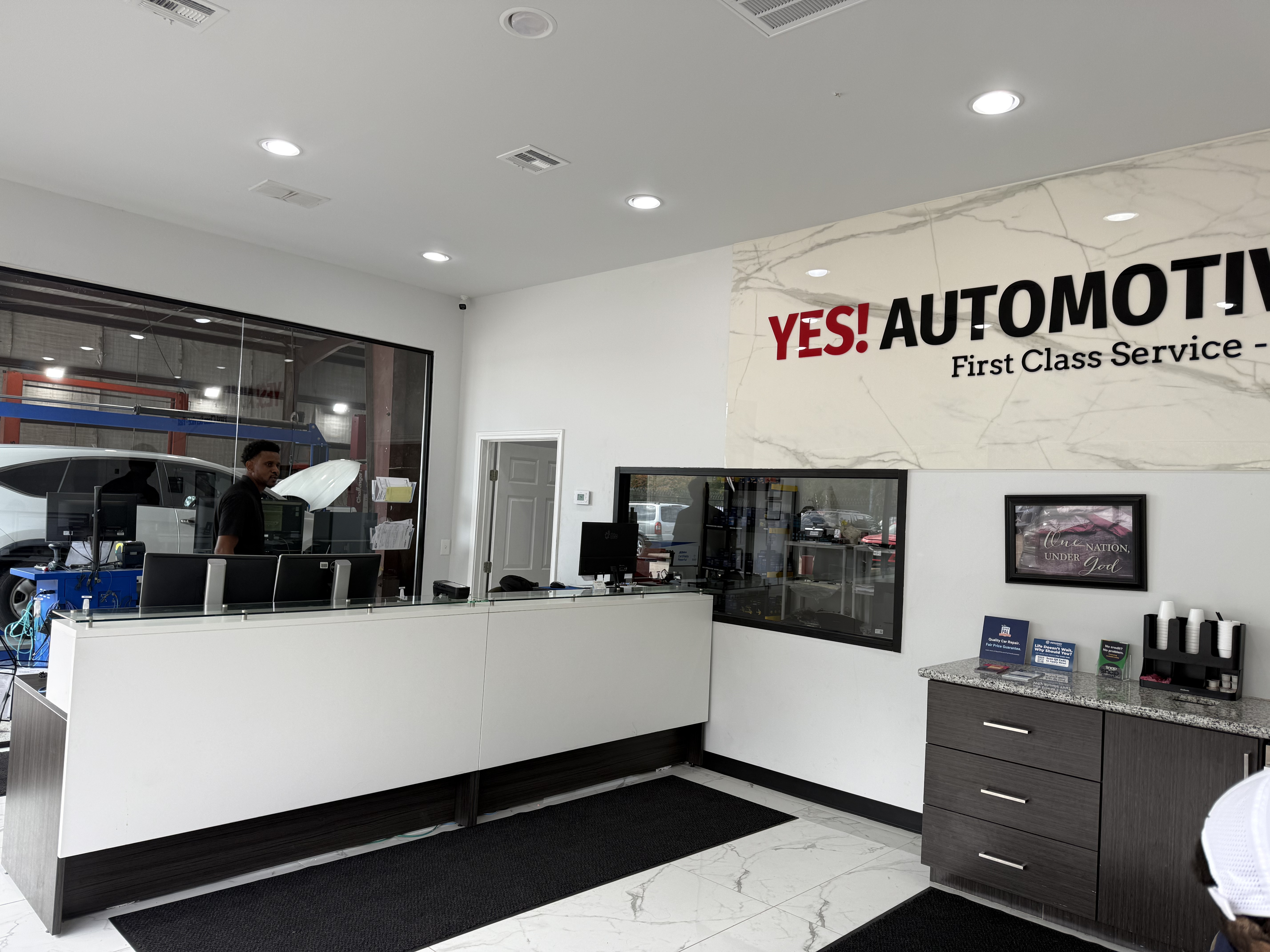 Interior view of YES! Automotive in Porter, TX, featuring a modern customer reception area, professional service advisor counter, prominent YES! Automotive branding, and a refreshment station. Large glass windows provide a view of the automotive repair bays.