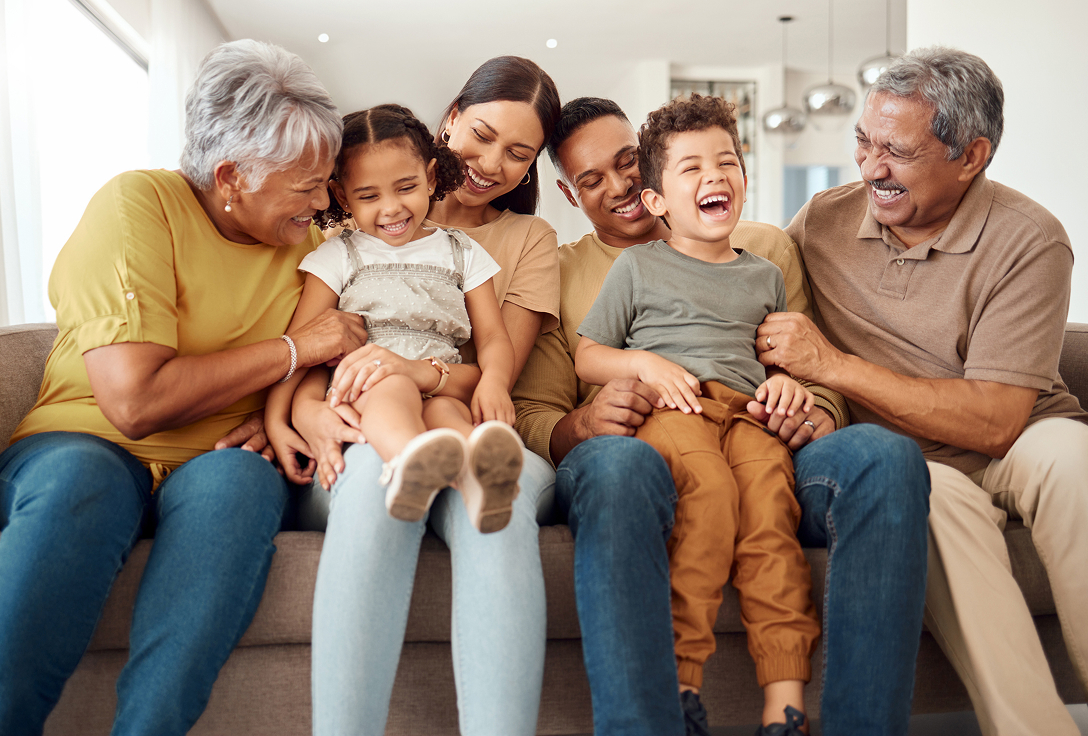 A joyful family sits on a couch, laughing together. Two kids are embraced by parents and grandparents. The room is bright, filled with warmth and happiness.