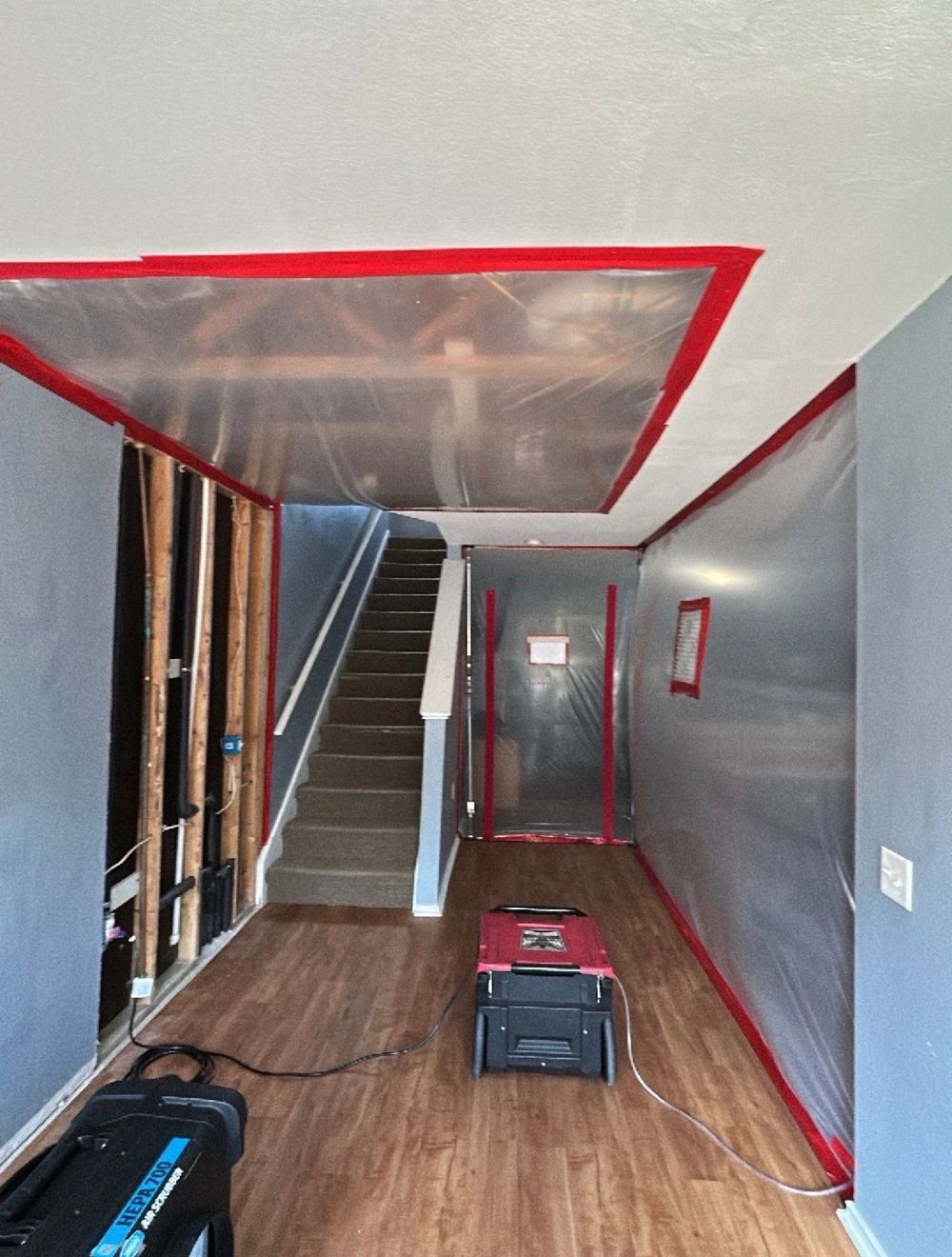 A residential hallway restoration area is enclosed with clear plastic sheeting and red tape across w 